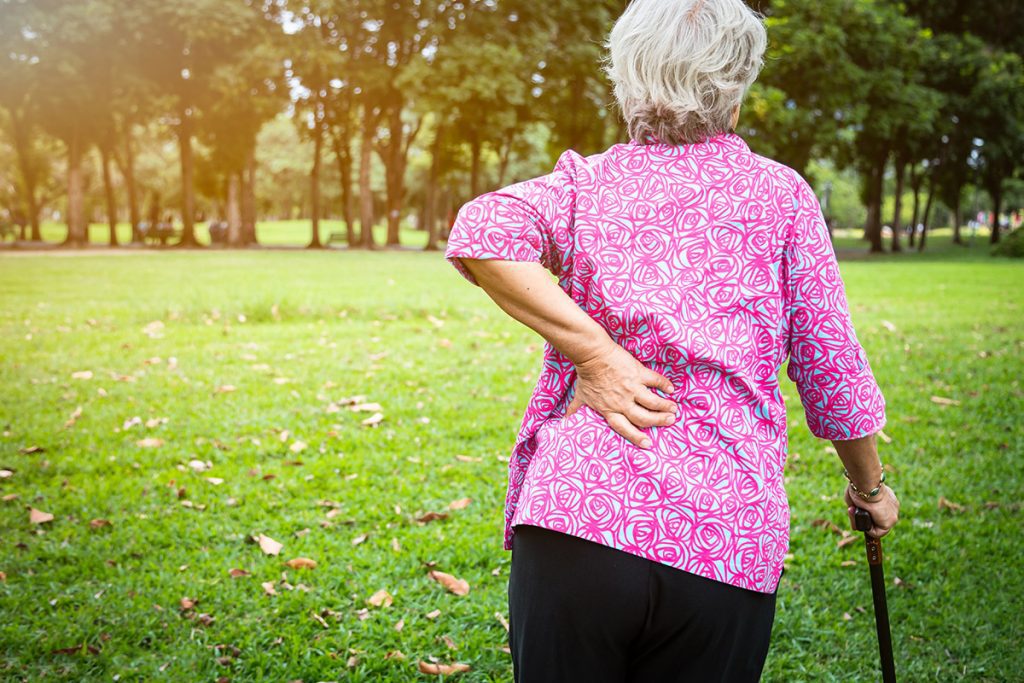 An elder woman holding the back of her hip in pain as she walks with a cane.