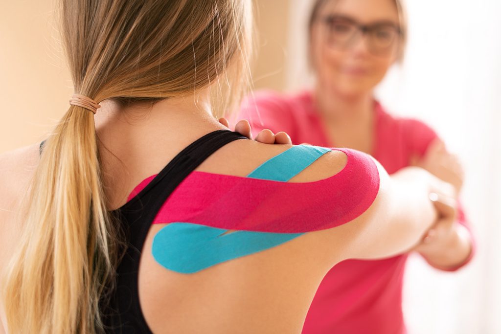 A patient wearing kinesio tape on her shoulder as her doctor helps her stretch.